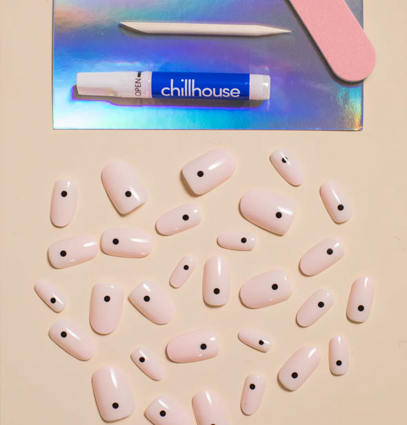 A smattering of whitish-pink press-on nails each with a single black dot next to an iridescent tray holding Chillhouse glue tube, pink nail file, and wooden cuticle pusher