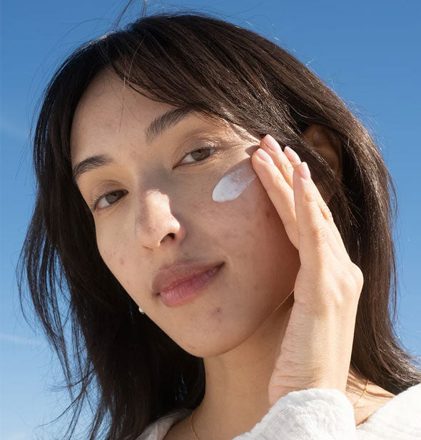 Model applies Coola Clear Skin Oil-Free Moisturizer to cheek against a blue sky backdrop