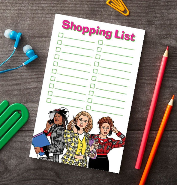 Clueless Shopping List notepad rests on a wooden surface with green and yellow paper clips, blue earbuds, and red and orange colored pencils