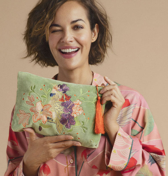 Winking and smiling model holds a green velvet pouch with embroidered cockatoo and flowers design