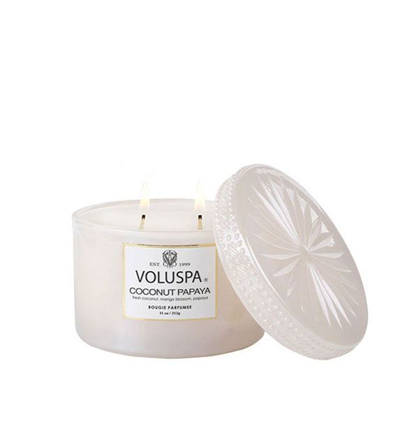 Lit two-wick Voluspa Coconut Papaya candle in frosted pearlescent glass jar with radially embossed matching lid removed and propped up alongside