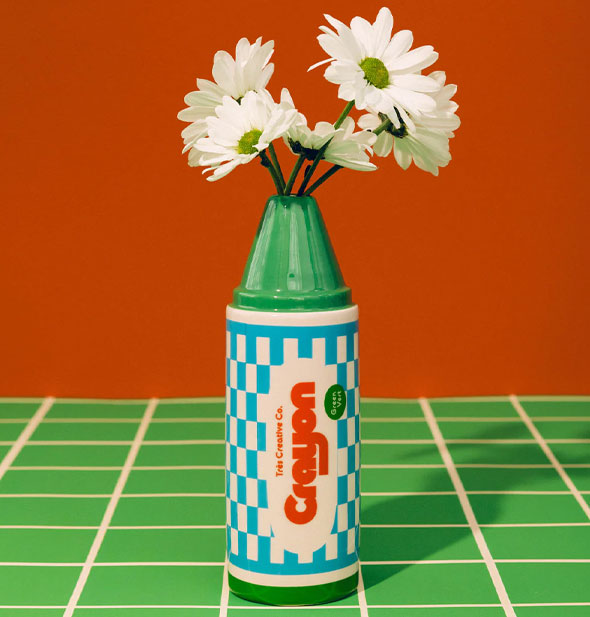 Crayon flower vase holds a small bunch of white daisies and rests on a green tile surface against a dark orange backdrop