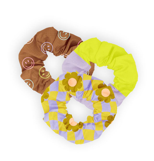 Set of three hair scrunchies: one brown with multicolored pastel smiley faces, one half periwinkle and half neon green, and one yellow and purple checker print with green and pink daisies