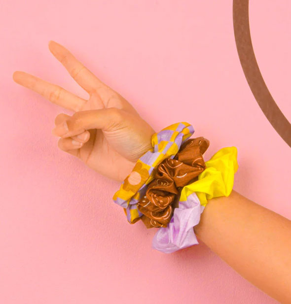 Model's outstretched arm with hand flashing a peace sign wears the three scrunchies from the Cool Funky Daisy set against a pink backdrop