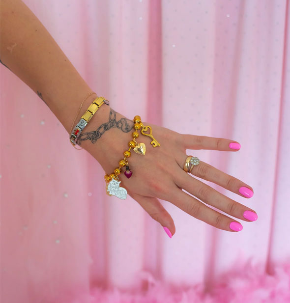 Model's outstretched arm wears a chunky gold ball chain charm bracelet against a pink draped backdrop