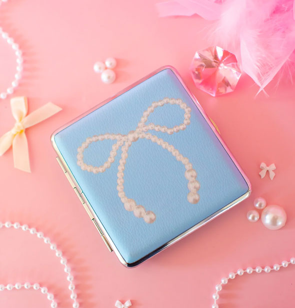 Rectangular blue cigarette case with white pearl bow graphic and silver frame on a pink surface scattered with pearls, bows, crystal, and feathers