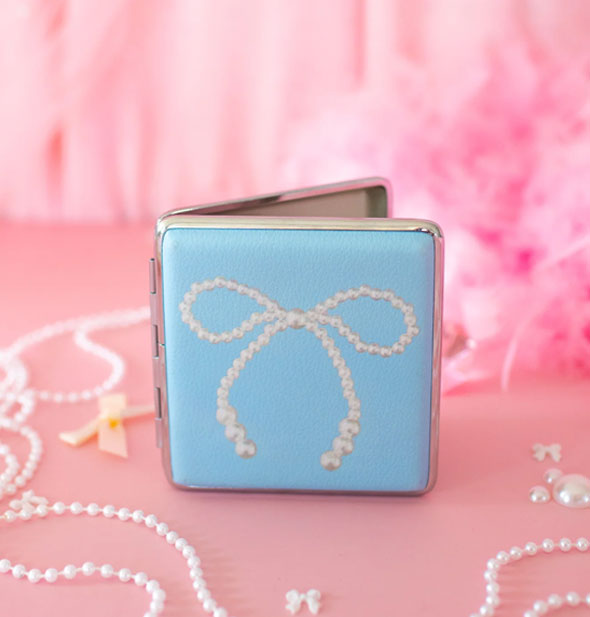 Opened rectangular blue cigarette case with white pearl bow graphic and silver frame on a pink surface scattered with pearls, bows, and feathers