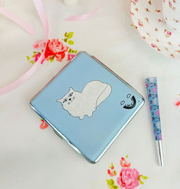 Rectangular blue cigarette case with white cat graphic and silver frame on a floral-patterned surface with a pre-rolled cigarette nearby