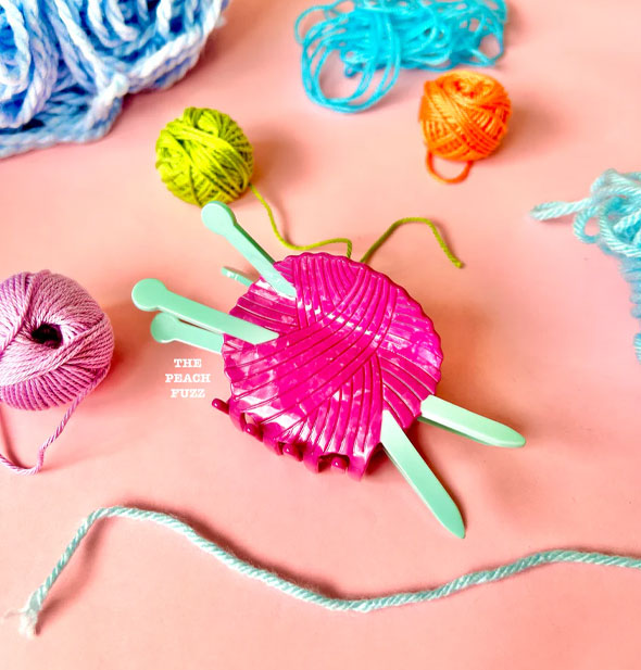 Hair clip designed as a pink ball of yarn pierced by two mint green knitting needles rests on a pink surface staged with multicolored balls and heaps of string and yarm