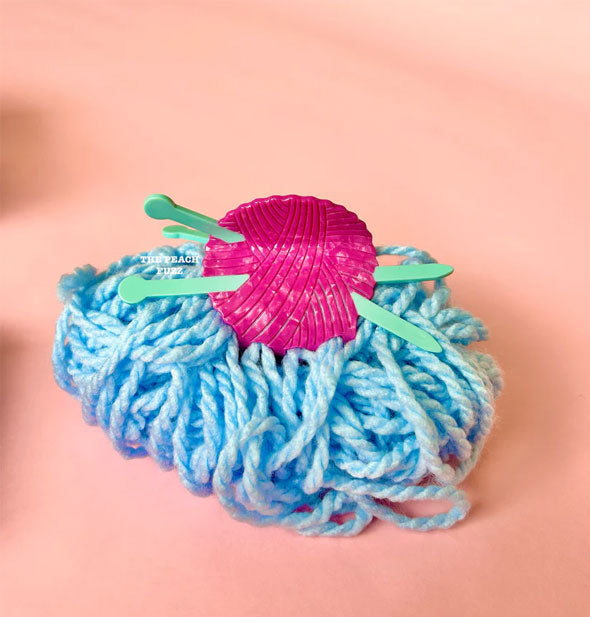 Hair clip designed to resemble a pink ball of yarn stuck with two green knitting needles rests on a bunch of blue yarn on a pink tabletop