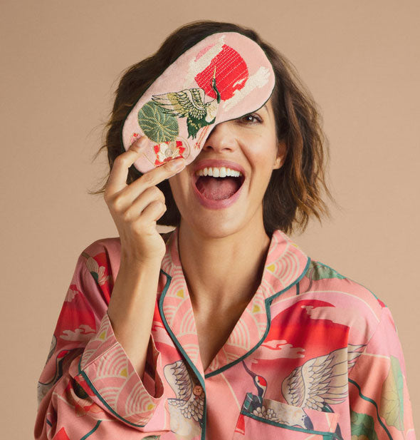 Smiling model playfully holds a Petal Pink Crane sleep mask over one eye