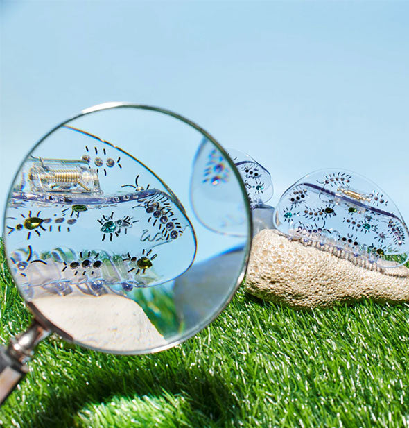Creepy Crawly Gem Claw Clips shown in a foreground magnifying glass and in the background on a rock on astroturf against a blue sky backdrop
