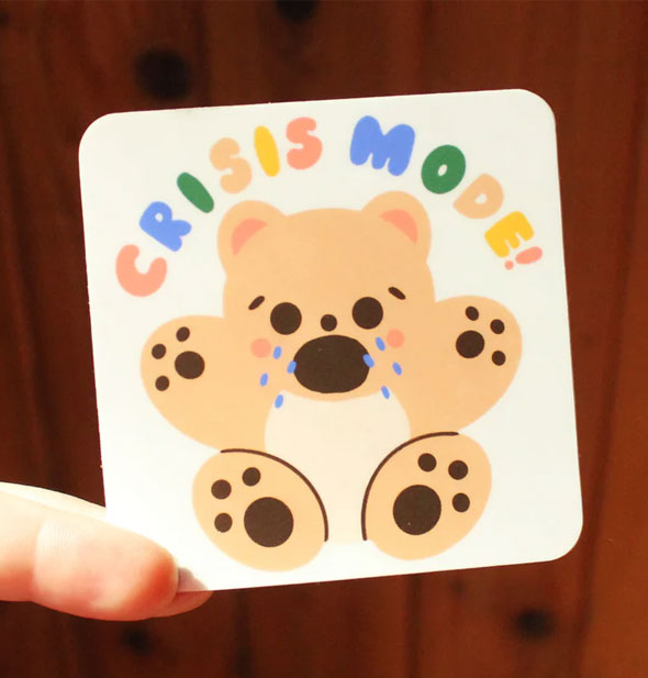 Crisis Mode bear sticker is pinched between a model's two fingers against a wooden backdrop