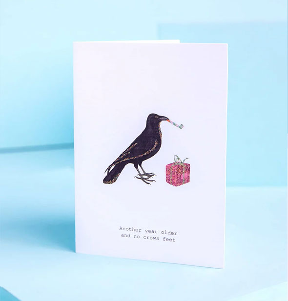 White greeting card on blue background features illustration of a black crow with party blower in its beak standing next to a pink wrapped gift says, "Another year older and no crows feet" at the bottom in fine print