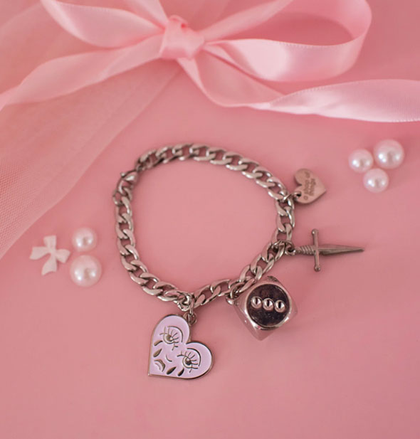 Thick silver curb chain with enamel crying heart, dice, dagger, engraved and silver heart charms is staged on a pink surface with white pearls and a pink satin bow
