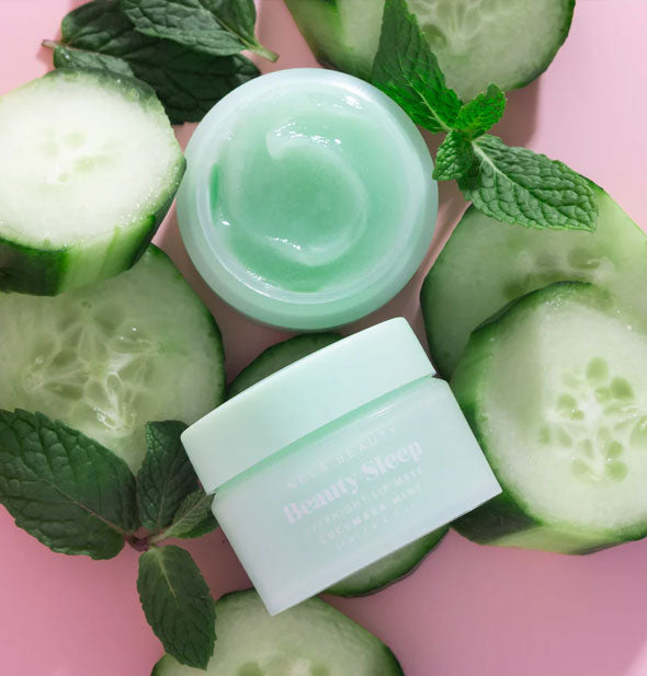 One closed and one opened pot of Cucumber Mint NCLA Beauty brand Beauty Sleep Overnight Lip Mask staged with slices of cucumber and sprigs of mint