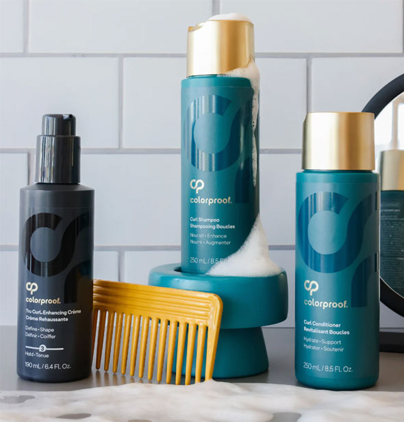 Bottles of ColorProof Tru Curl Enhancing Crème, Curl Shampoo, and Curl Conditioner rest on a bathroom countertop with sudsy lather, a gold wide tooth comb, and mirror in the background against a tiled wall