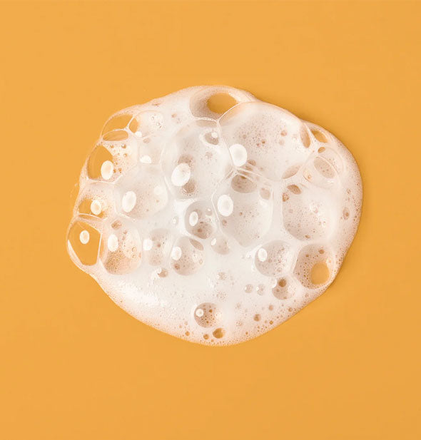 Closeup of white, bubbly Verb Curl Foaming Gel on an orange surface