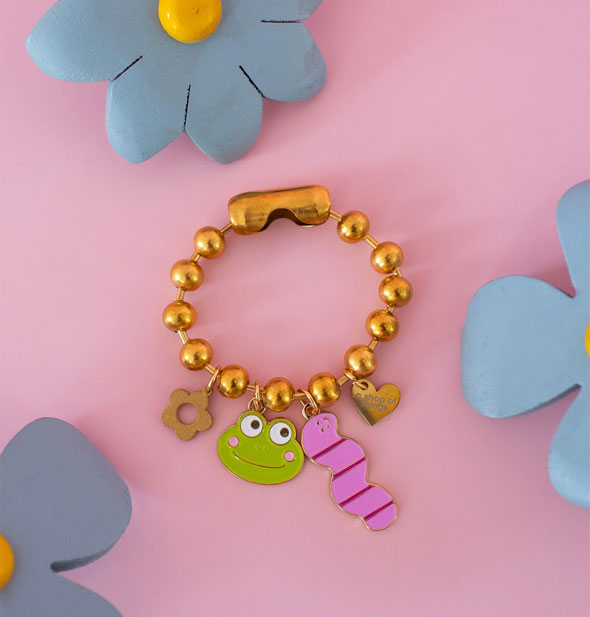 Chunky gold ball chain accessory with gold daisy, green enamel frog face, pink worm, and gold heart charms attached; surrounding are painted wooden flowers on a pink surface