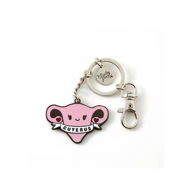 Pink enamel keychain charm with silver hardware attached resembles a smiling cartoon uterus with hearts on the ends of its fallopian tubes, wrapped in a white banner that says, "Cuterus"