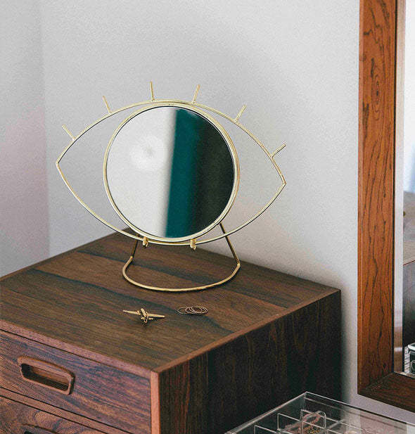 Gold-framed eye-shaped makeup mirror staged on a wooden cabinet top with accessories