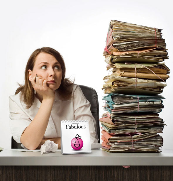An overwhelmed employee stares at a large pile of messy files on her desk next to a Daily Mood flip book that says, "Fabulous" with pink smiley face illustration