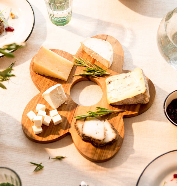Wooden daisy-shaped serving board holds a variety of cheeses with fresh herbs on a staged tabletop