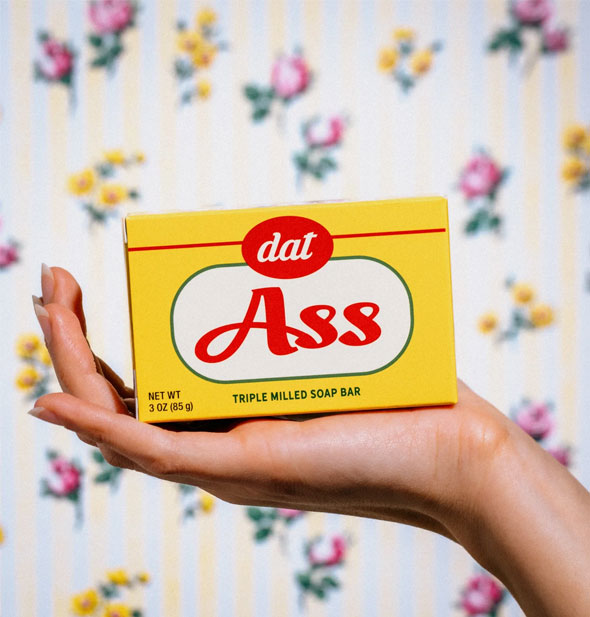 Model's hand holds a yellow, white, and red box of Dat Ass Triple Milled Bar Soap up in front of a floral backdrop