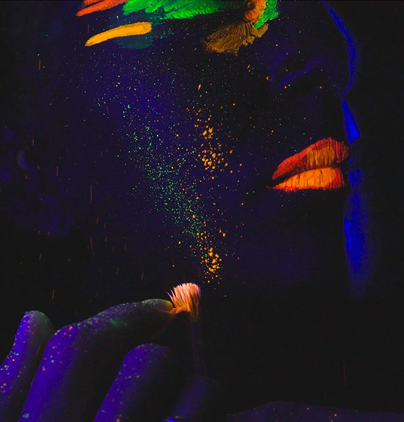 Model wears neon face paint in streaks and splatters that glow under a blacklight