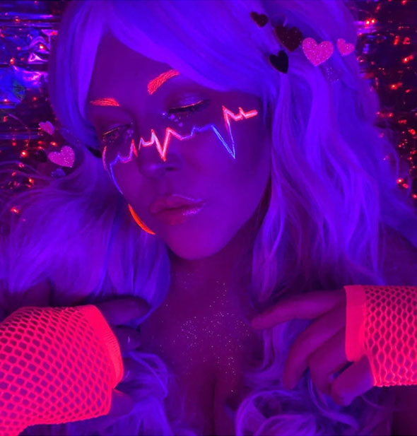 Model wears DayGlo face paint including that which resembles a heart monitor line tracing that glows pink and purple under a blacklight
