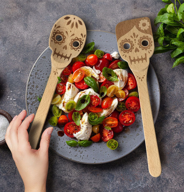 Model reaches for the slotted sugar skull spoon that rests on the edge of a salad platter next to its matching sugar skull spatula