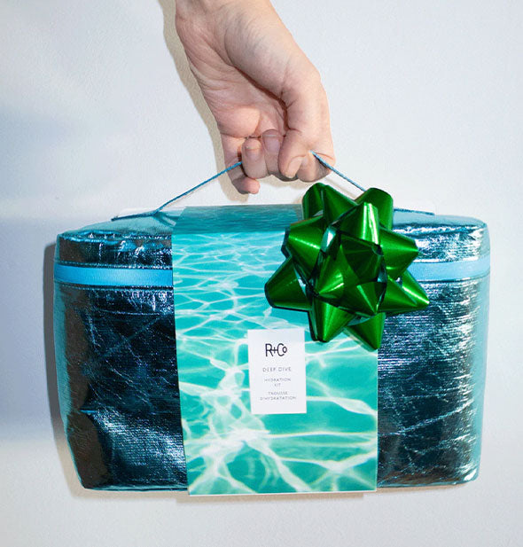 Model's hand holds a metallic blue R+Co Deep Dive kit bag with metallic green ribbon attached by its handle