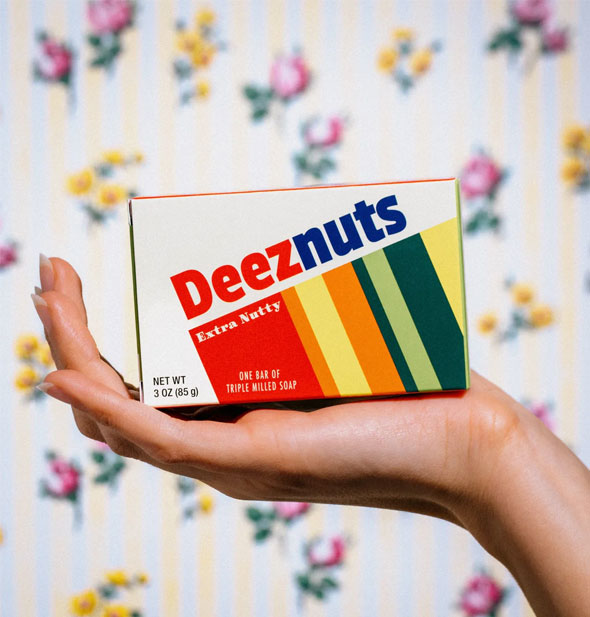Model's hand holds a box of Deez Nuts bar soap with bright color bands design up in front of a floral backdrop