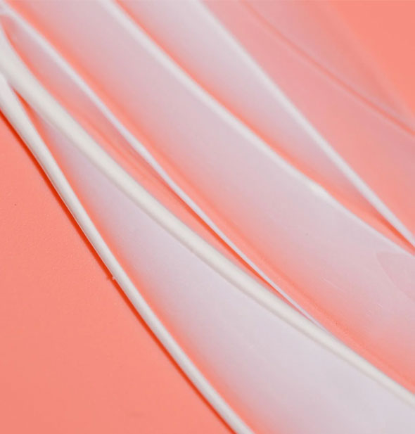 Closeup of sheer, whitish streaks of Coola Dew Good Illuminating Serum Sunscreen on a peach-colored surface