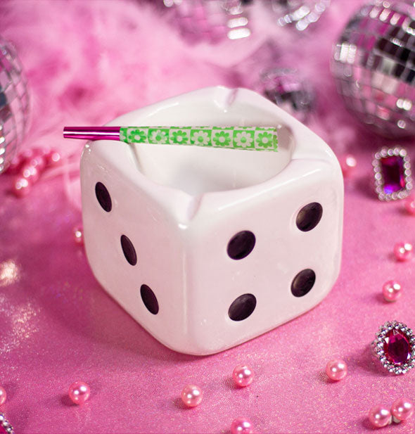 White ceramic cube-shaped ashtray designed to resemble a playing die with black "pips" holds a green and pink rolled cigarette and rests on a pink surface sprinkled with pink pearls, gemstones, and disco balls