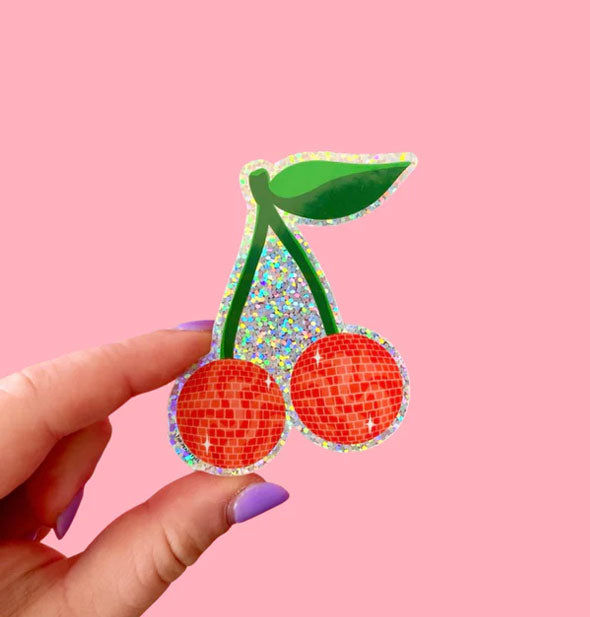 Model's hand holds a sticker with design of two red cherries which resemble disco balls on a green stem with leaf on a glittery holographic background