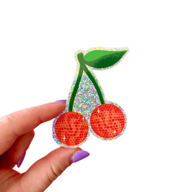 Model's hand holds a sticker with design of two red cherries which resemble disco balls on a green stem with leaf on a glittery holographic background