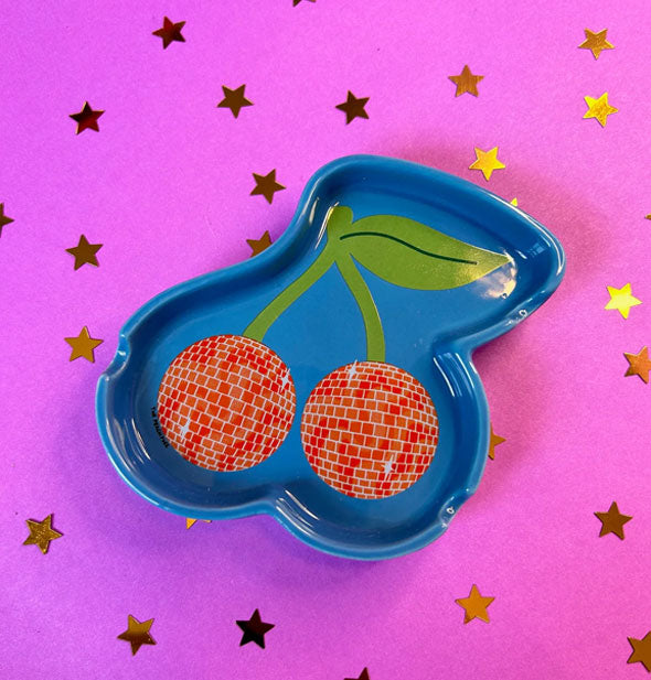 Blue ashtray with red and green disco ball cherries design on a purple surface scattered with gold star confetti