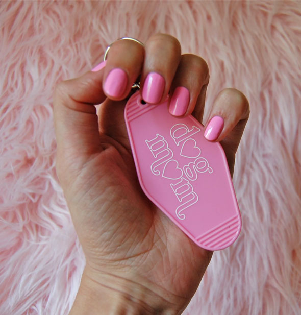 Model's hand with pink fingernails clutches a pink Dog Mom motel-style keychain tab against a pink fur backdrop