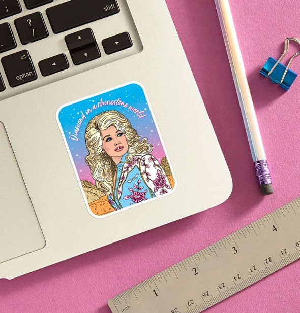 Dolly Parton "Diamond in a rhinestone world" sticker is applied to a laptop base panel and staged with pencil, ruler, and blue binder clip on a pink surface