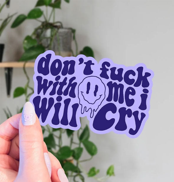 Model's hand holds a purple sticker that says, "Don't fuck with me I will cry" in warped lettering surrounding a melting smiley face graphic