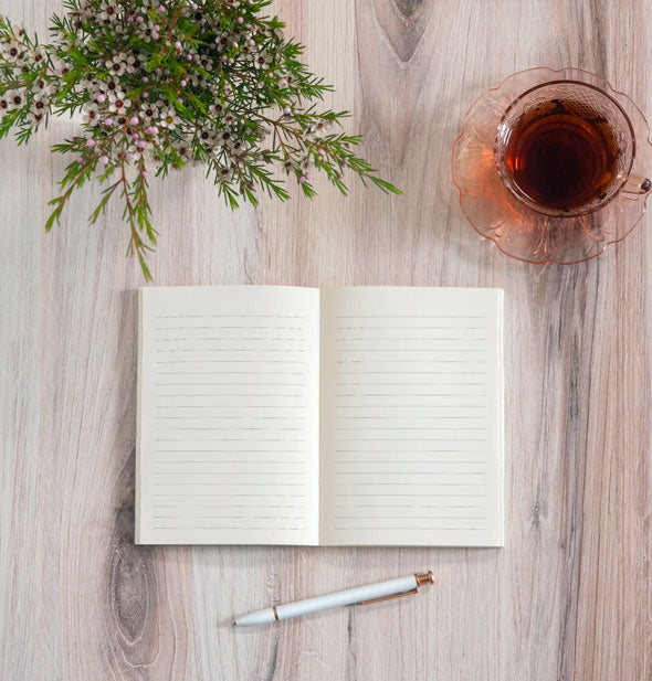 Opened lined notebook with pen on a wooden surface staged with a cup of tea and floral arrangement