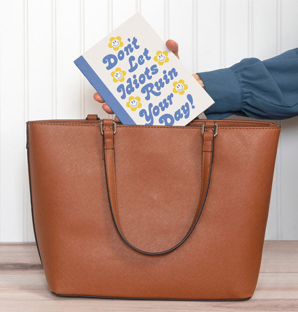 Model pulls a Don't Let Idiots Ruin Your Day journal from a brown handbag