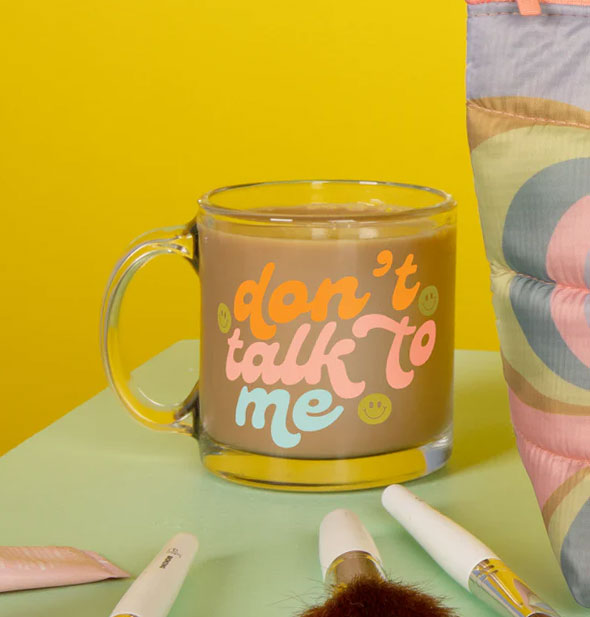 Don't Talk to Me glass mug filled with a coffee-like beverage rests on a mint green tabletop with a puffy bag and noisemakers against a mustard yellow backdrop