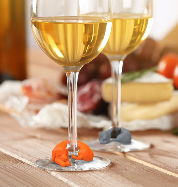 Orange and gray silicone dachshund drink markers wrap around the stems of two wine glasses on a tabletop