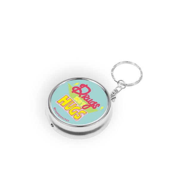 Round silver pill case with keychain attachment says, "Drugs not hugs" in pink and yellow lettering on a blue background