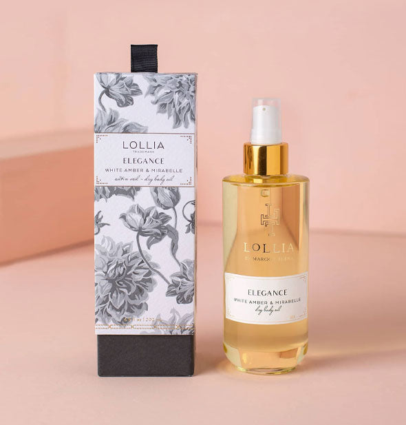 Bottle of golden Lollia Elegance White Amber & Mirabelle Dry Body Oil next to box featuring black and white floral motif, both set against a pink backdrop