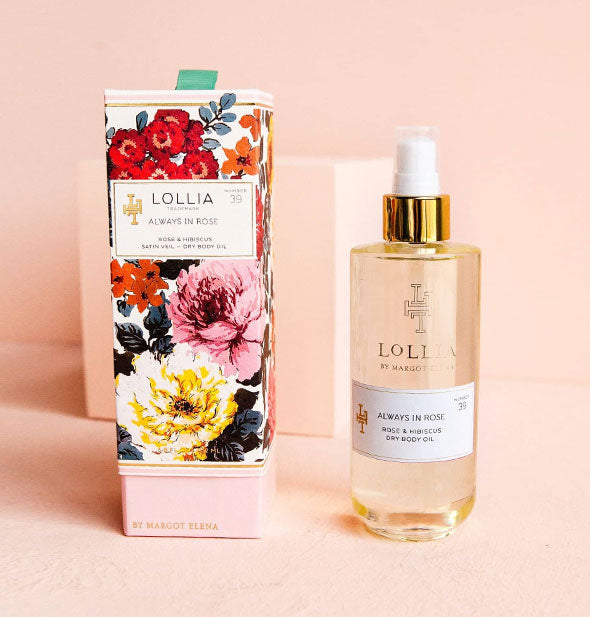 Cylindrical glass bottle of Lollia Always In Rose Dry Body Oil accented with gold and white next to its colorful floral-print box on a pink backdrop