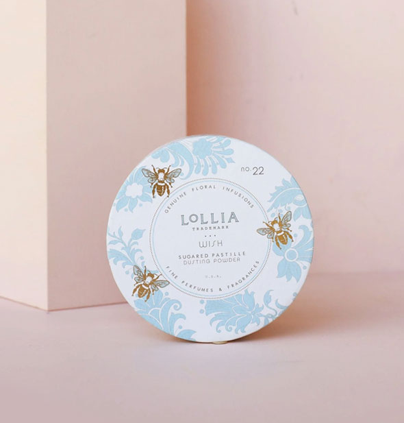 Round white container of Lollia Wish Sugared Pastille Dusting Powder features a design of blue florals and gold bees