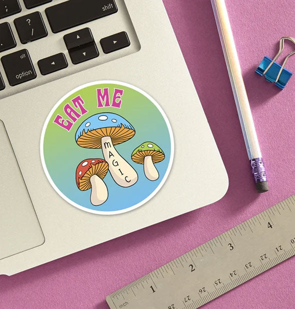 Round "Eat Me" magic mushrooms sticker is applied to a laptop base panel and staged with pencil. ruler, and blue binder clip on a pink surface